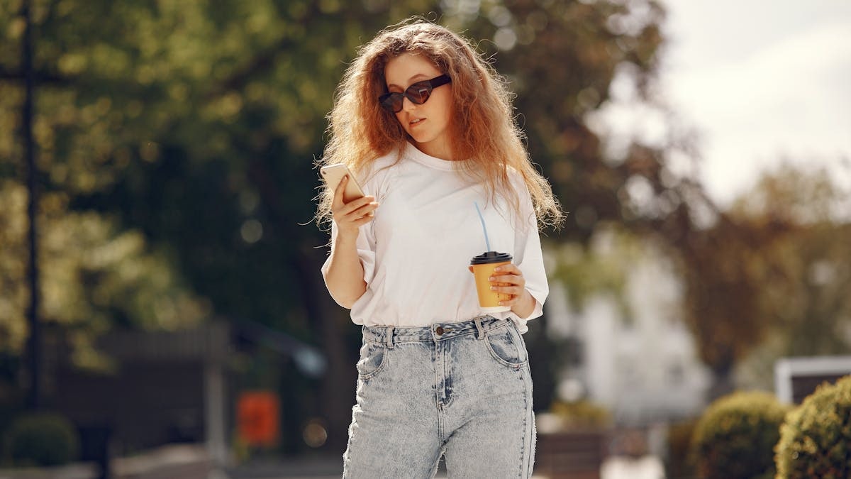 Stylish woman with sunglasses enjoys coffee while using smartphone outdoors.